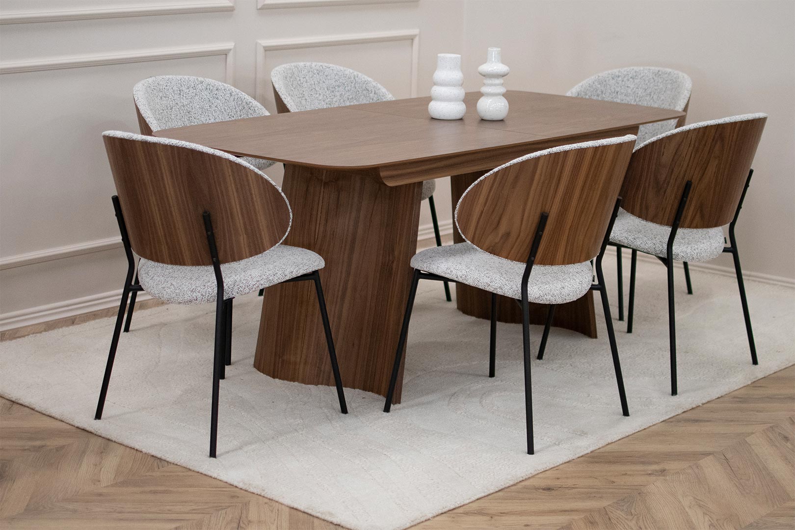 Трпезариски сет со маса Сефра од орев со две направилна форма дрвени ногарки, со столици Алексис со фурниран грб орев и светло крем мебел штоф, поставени на волнен килим во бела боја.