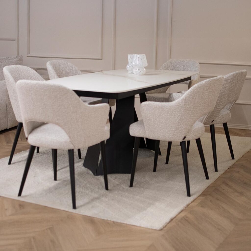 Трпезариска маса со керамичка плотна во бела боја со крем шари поставена на две црни метални ногарки во комбинација со столици со раконаслон во бел мебел штоф со црни дрвени ногарки.