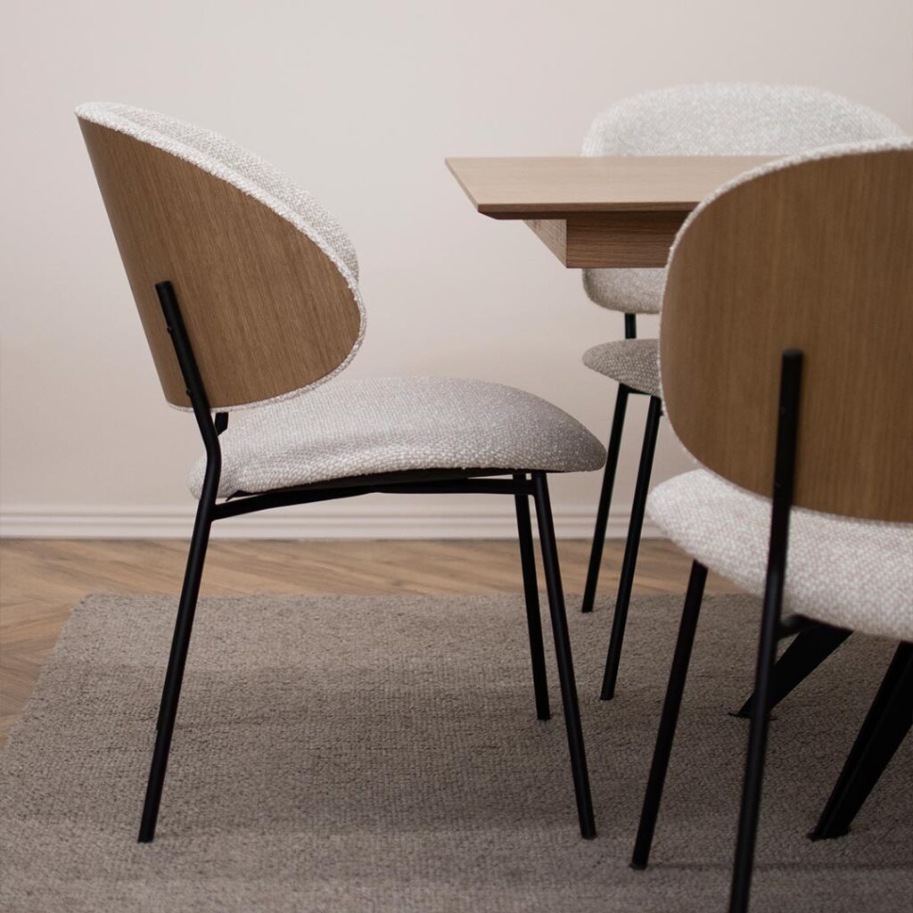 Трпезариска столица, со удобно седло во беж мебел штоф и фурнир од даб во грбот, поставена на црни елегантни метални ногарки.