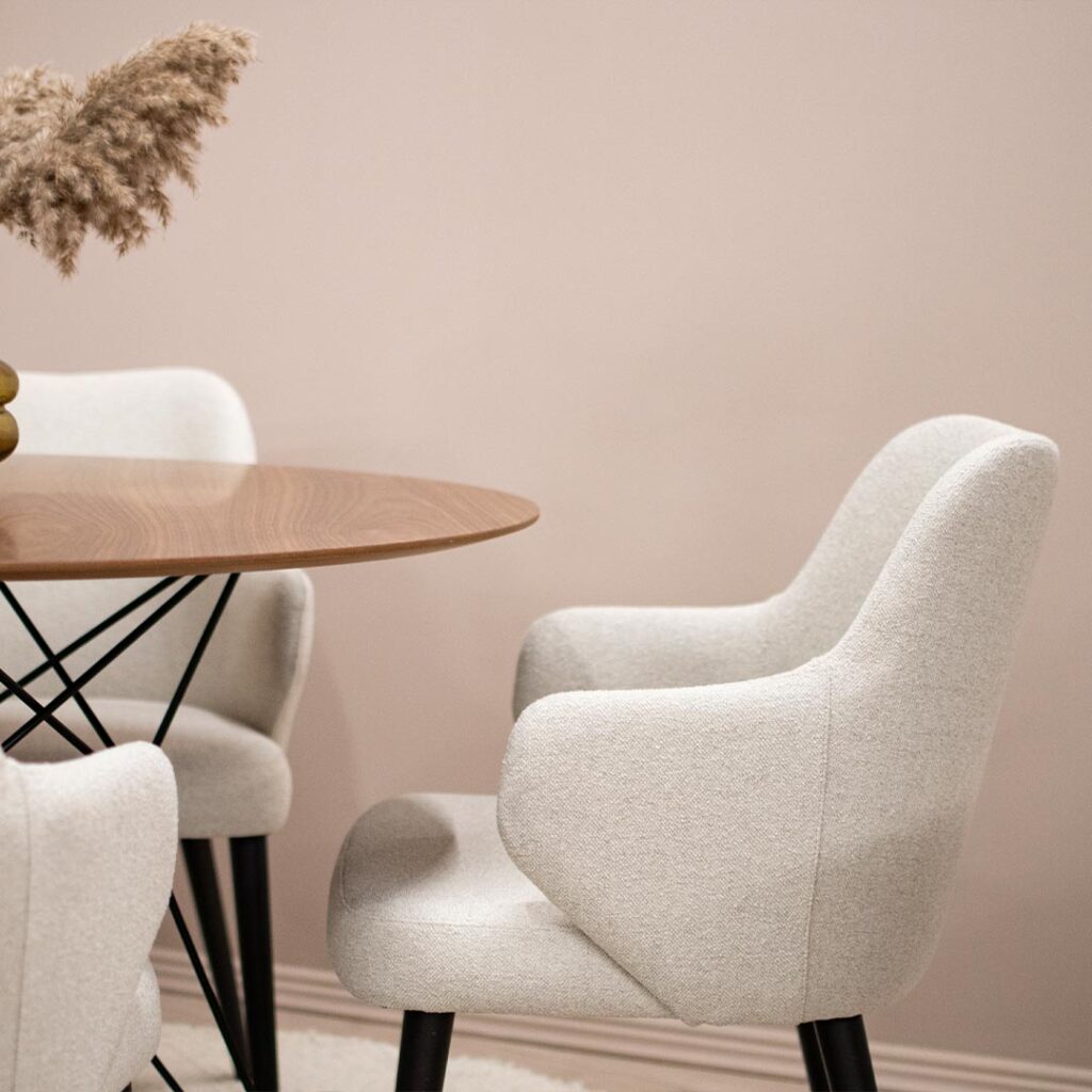 Трпезариска столица со раконаслон во крем мебл штоф и црни ногарки поставена на мала кружна маса со орев плотна.