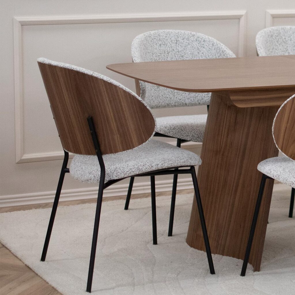 Трпезариски сет со маса Сефра од орев со две направилна форма дрвени ногарки, со столици Алексис со фурниран грб орев и светло крем мебел штоф, поставени на волнен килим во бела боја.