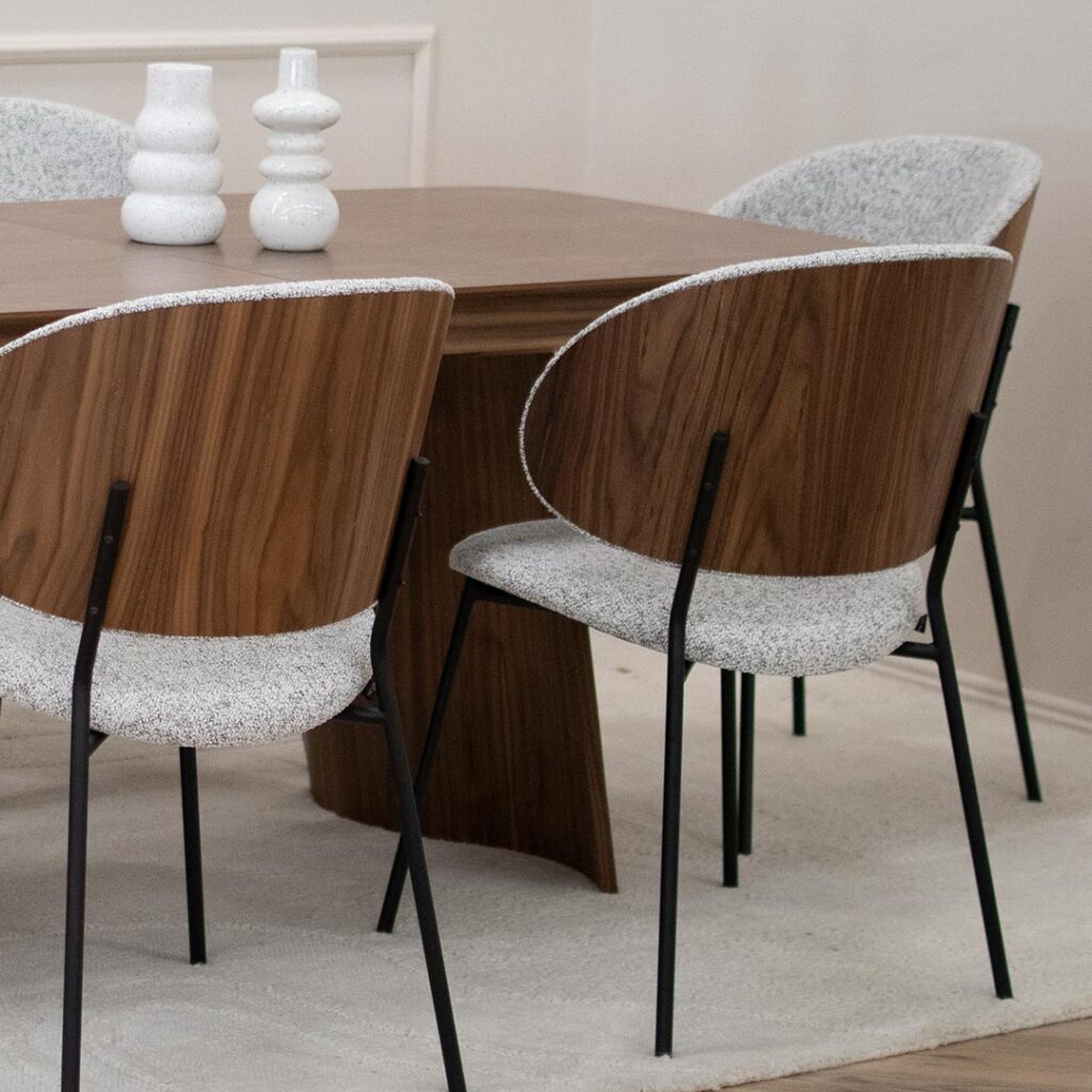 Трпезариски сет со маса Сефра од орев со две направилна форма дрвени ногарки, со столици Алексис со фурниран грб орев и светло крем мебел штоф, поставени на волнен килим во бела боја.