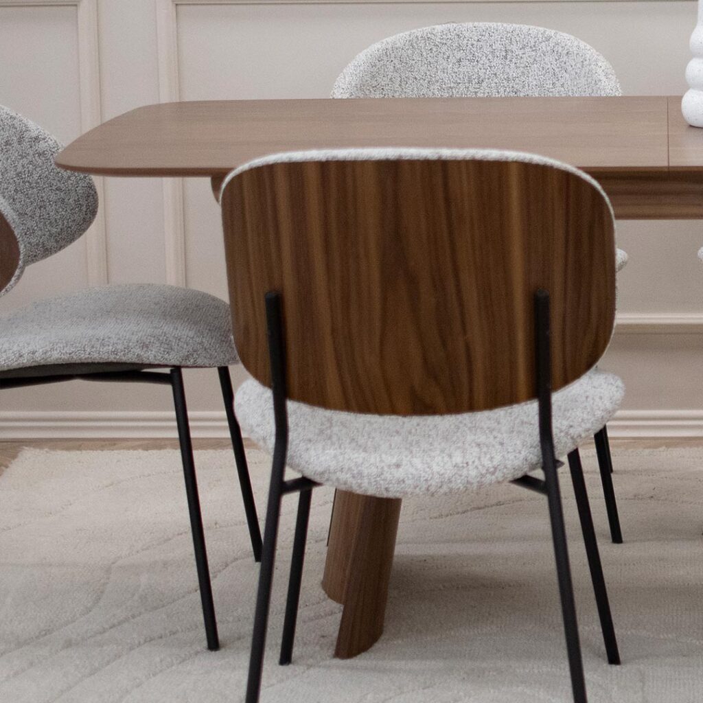 Трпезариски сет со маса Сефра од орев со две направилна форма дрвени ногарки, со столици Алексис со фурниран грб орев и светло крем мебел штоф, поставени на волнен килим во бела боја.