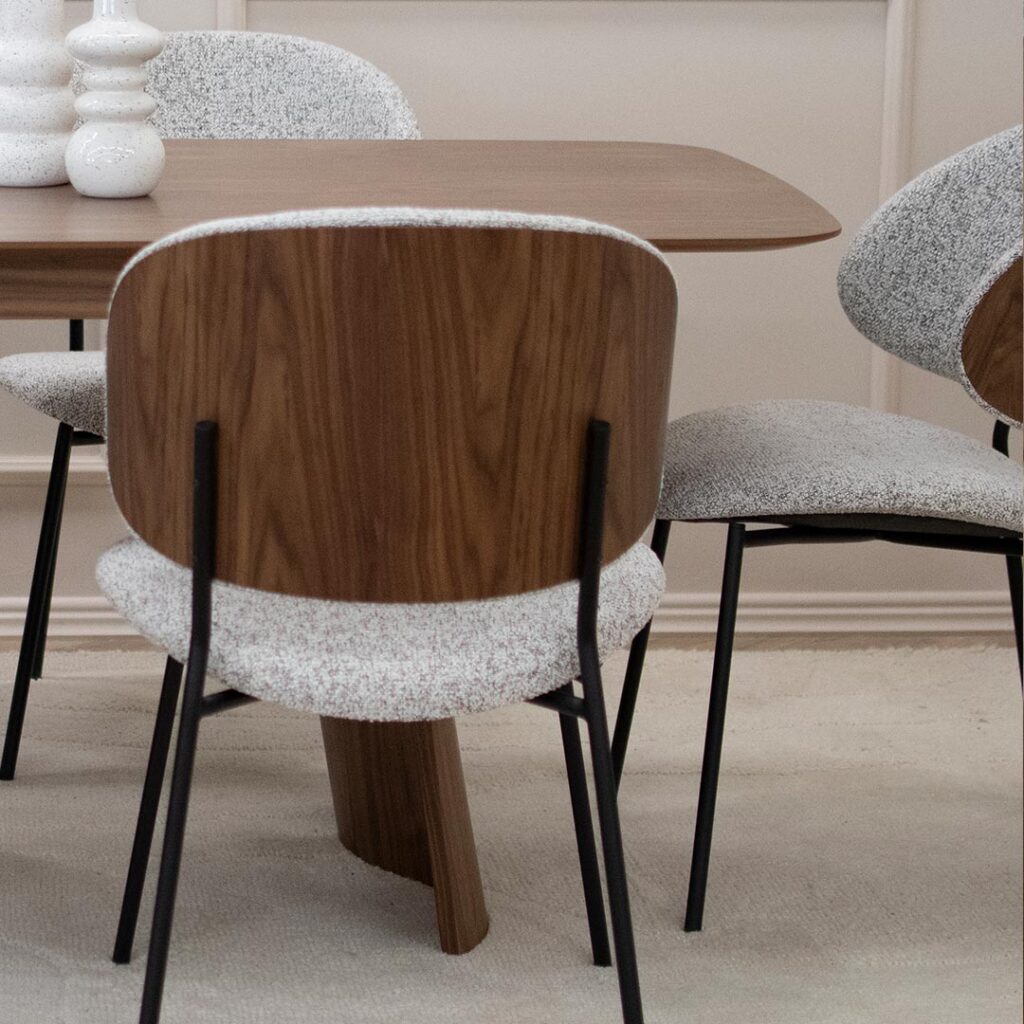 Трпезариски сет со маса Сефра од орев со две направилна форма дрвени ногарки, со столици Алексис со фурниран грб орев и светло крем мебел штоф, поставени на волнен килим во бела боја.