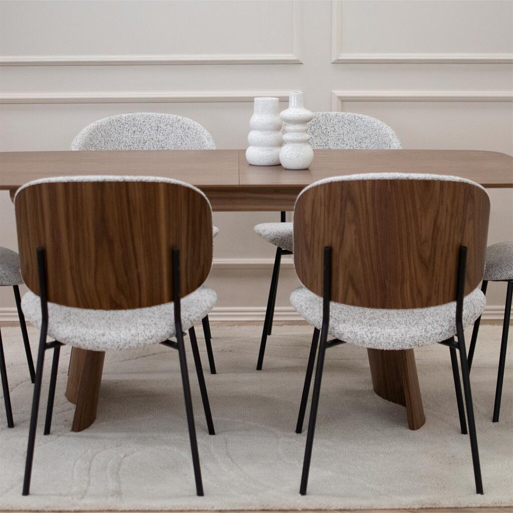 Трпезариски сет со маса Сефра од орев со две направилна форма дрвени ногарки, со столици Алексис со фурниран грб орев и светло крем мебел штоф, поставени на волнен килим во бела боја.