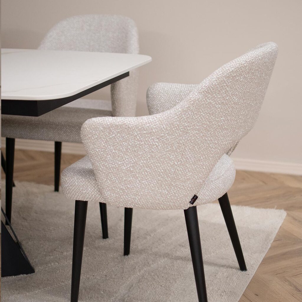 Трпезариска маса со керамичка плотна во бела боја со крем шари поставена на две црни метални ногарки во комбинација со столици со раконаслон во бел мебел штоф со црни дрвени ногарки.