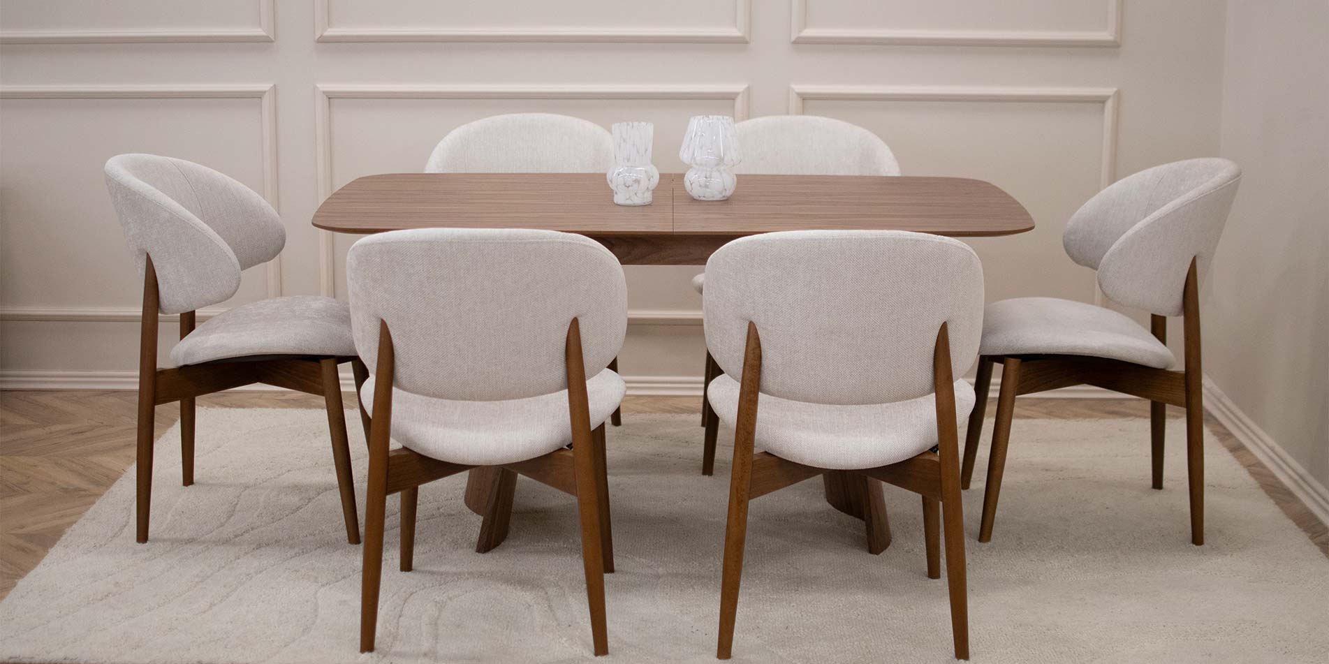 Трпезариска столица Celine со бука ногарки со светло крем мебел штоф, поставени на маса од дрво орев.