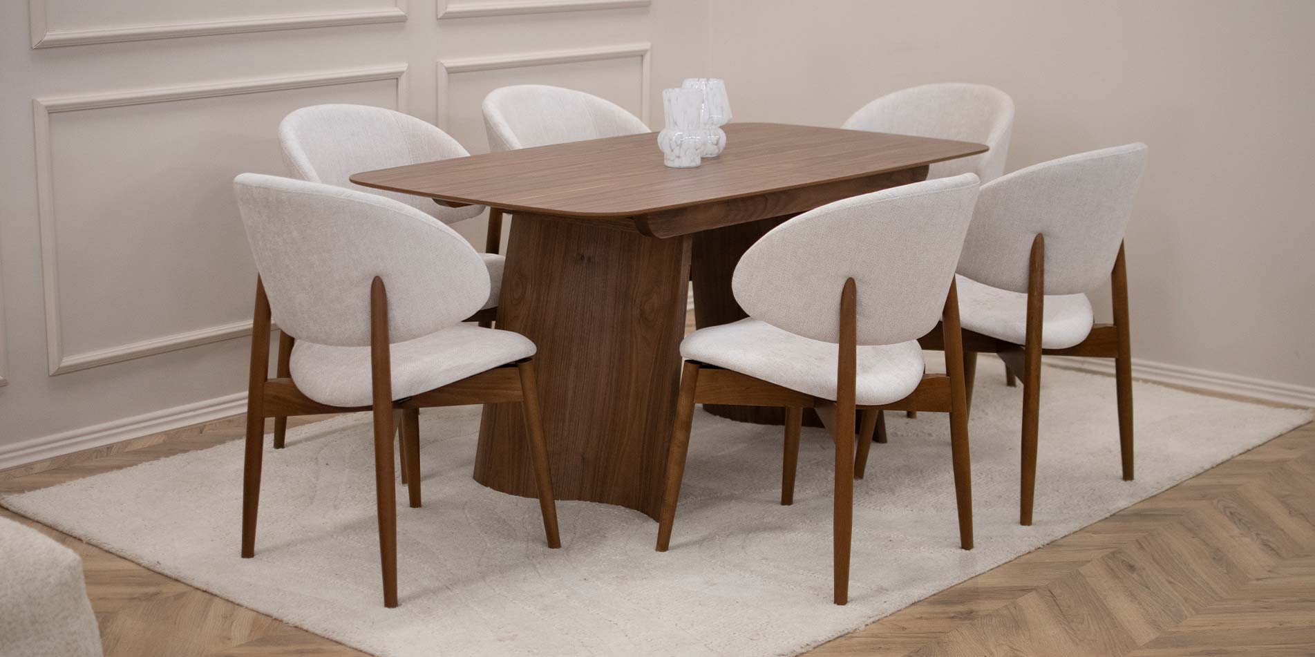 Трпезариска столица Celine со бука ногарки со светло крем мебел штоф, поставени на маса од дрво орев.